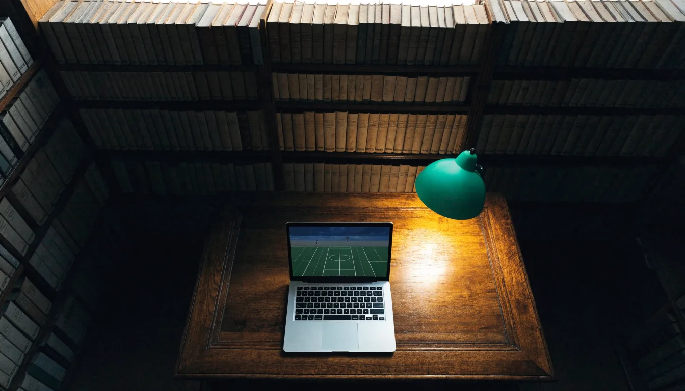 Eine Bibliothek mit hohen Holzregalen voller Bücher, im Vordergrund ein Lesetisch mit grüner Lampe.
