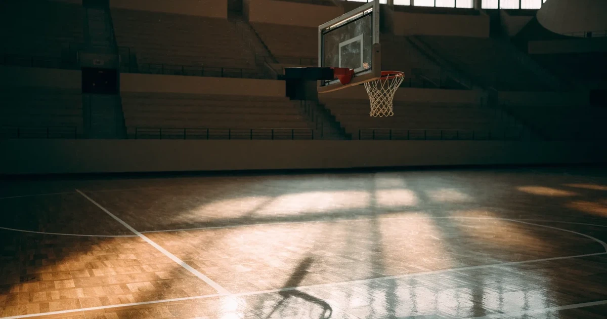 Ein Basketballkorb mit Netz in einer leeren Halle, Licht fällt von hohen Fenstern auf den glänzenden Parkettboden.