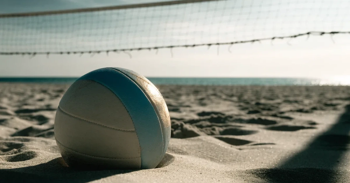 Ein Volleyball liegt auf feinem hellem Sand, ein gespanntes Netz verschwimmt dahinter, das Licht kommt von einem weiten Meer.
