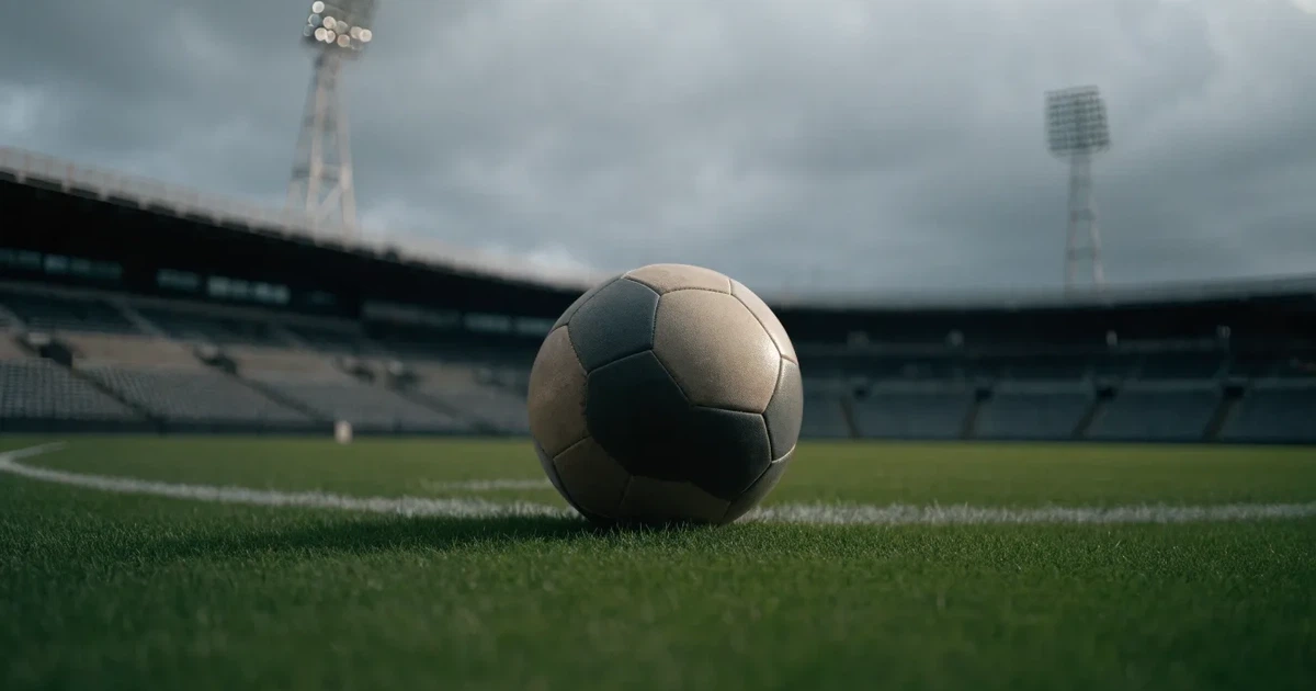 Ein Fussball liegt auf dem Anstosspunkt eines frisch gemähten Rasens, dahinter eine grosse Tribüne unter bewölktem Himmel