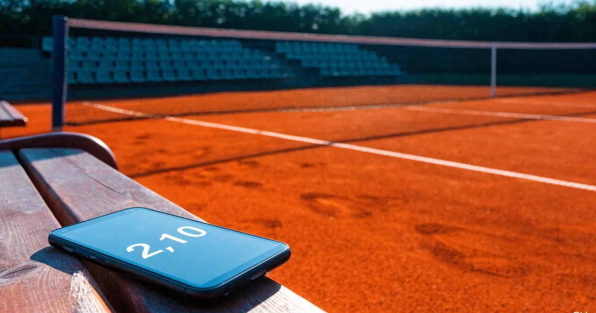 Ein Tennisplatz aus rotem Sand liegt im ersten Licht des Morgens, das Netz hängt still und die Linien sind frisch gezogen.