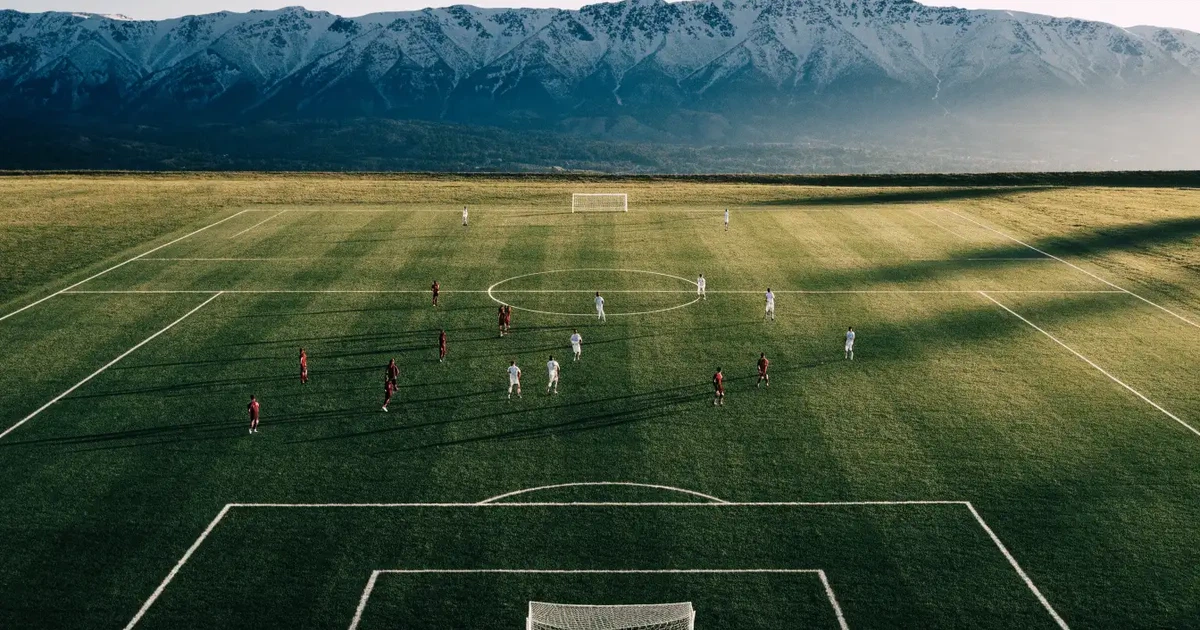 Ein Fussballplatz aus der Vogelperspektive mit sauberen Linien auf dichtem Rasen, in der Ferne verschneite Berge.