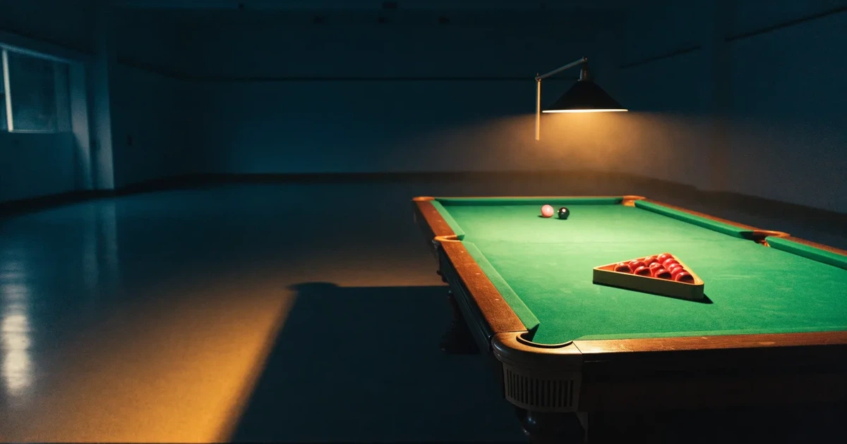 Ein Snookertisch mit grünem Tuch und einem Dreieck aus roten Kugeln unter einer tief hängenden Lampe in einer leeren Halle.