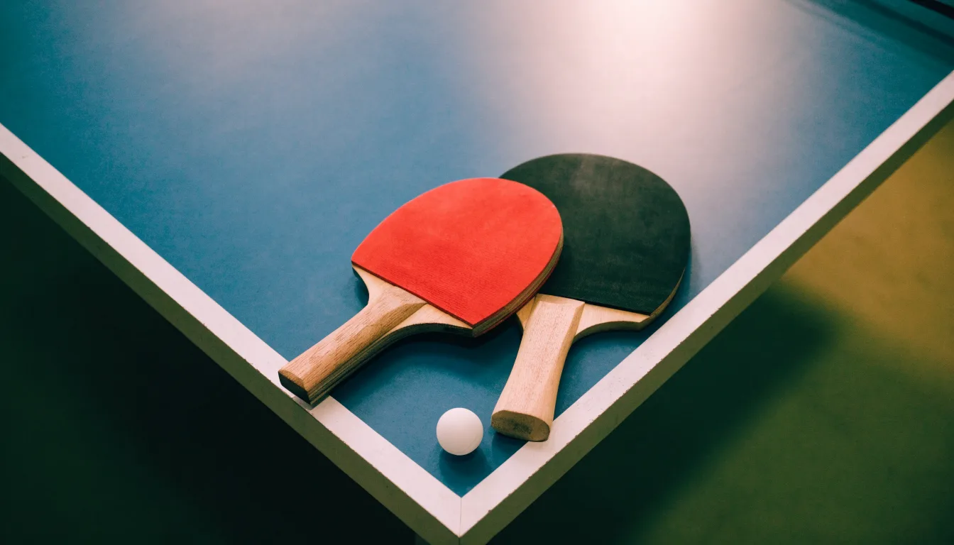 Zwei Tischtennisschläger liegen gekreuzt am Rand einer Platte, daneben ein kleiner weisser Ball.