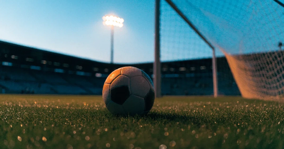 Ein Fussball liegt auf dem Anstosspunkt eines leeren Rasenplatzes, dahinter ragen die Ränge eines grossen Stadions in der Abenddämmerung auf.